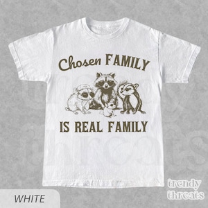 May include: White t-shirt with a brown graphic of a raccoon, a squirrel, a mouse, and a rat wearing sunglasses. The text "Chosen Family is Real Family" is printed on the shirt.
