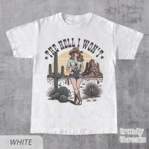 May include: White t-shirt with a graphic of a woman in a cowboy hat and denim shorts standing in a desert landscape with a cactus and the text "The Hell I Won't".