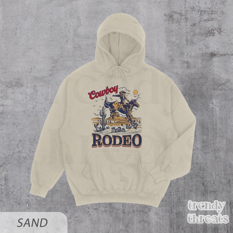 May include: A sand-colored hoodie with a vintage-style illustration of a cowboy riding a horse in a desert landscape. The text "Cowboy Rodeo" is printed on the hoodie.