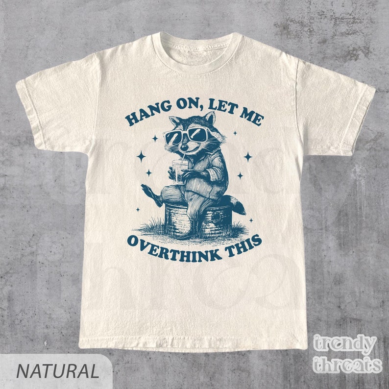 May include: A white t-shirt with a blue illustration of a raccoon wearing sunglasses and drinking from a can. The text "Hang on, let me overthink this" is printed on the shirt.