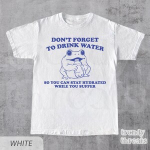 K&ouml;nnte beinhalten: Wei&szlig;es T-Shirt mit einem blauen Comic-Frosch, der eine Wasserflasche h&auml;lt. Der Text auf dem Shirt lautet "Vergiss nicht, Wasser zu trinken, damit du hydriert bleibst, w&auml;hrend du leidest."