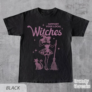 Flat lay black color shirt with quote 'Support Your Local Witches' and retro Halloween witch graphic.
