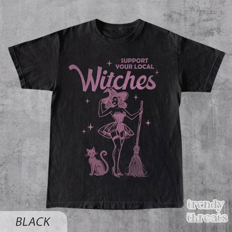 Flat lay black color shirt with quote 'Support Your Local Witches' and retro Halloween witch graphic.