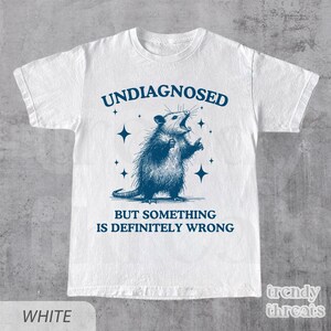 May include: White t-shirt with a blue illustration of an opossum with the text "UNDIAGNOSED BUT SOMETHING IS DEFINITELY WRONG". The opossum is drawn with its mouth open and arms raised.
