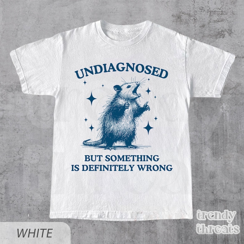 May include: White t-shirt with a blue illustration of an opossum with the text "UNDIAGNOSED BUT SOMETHING IS DEFINITELY WRONG". The opossum is drawn with its mouth open and arms raised.