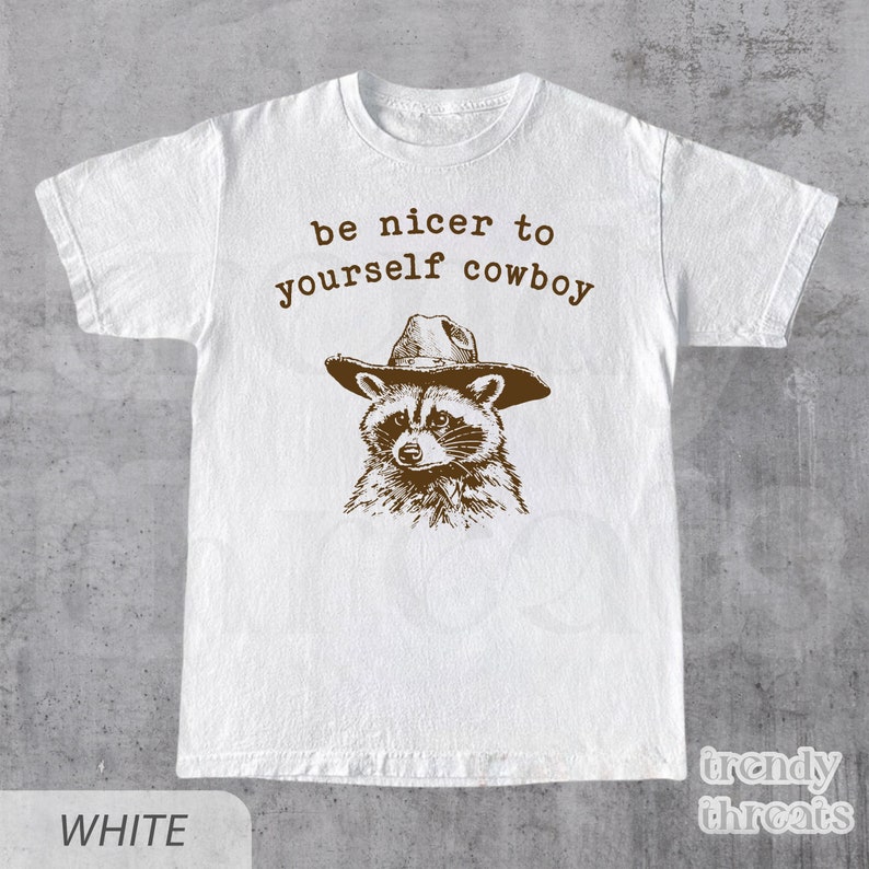 May include: White t-shirt with a brown illustration of a raccoon wearing a cowboy hat. The text "be nicer to yourself cowboy" is printed above the raccoon.