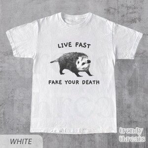 May include: White t-shirt with a black and white illustration of a possum and the text "Live Fast Fake Your Death."