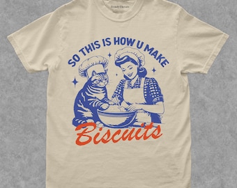May include: Cream-colored t-shirt featuring a blue and red graphic. The graphic depicts a woman and a cat wearing chef hats, mixing ingredients in a bowl. Text reads "SO THIS IS HOW U MAKE Biscuits."