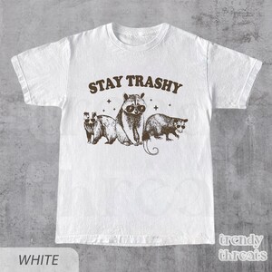 May include: White t-shirt with a brown graphic of raccoons and opossums wearing sunglasses. The text "STAY TRASHY" is above the animals.