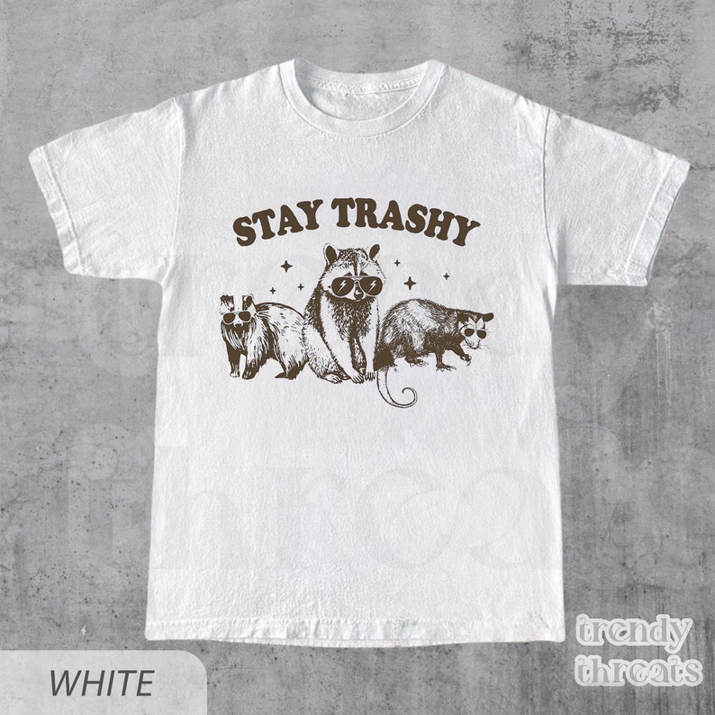 May include: White t-shirt with a brown graphic of raccoons and opossums wearing sunglasses. The text "STAY TRASHY" is above the animals.