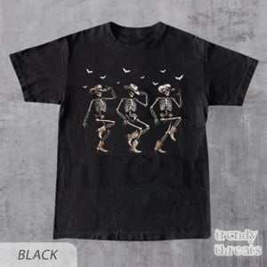 May include: Black t-shirt with a graphic of three skeletons wearing cowboy hats and boots, dancing. The skeletons are outlined in white and have a vintage look. Bats are flying above the skeletons.