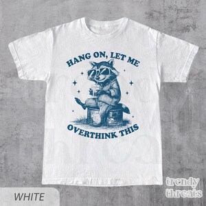 May include: White t-shirt with a blue graphic of a raccoon wearing sunglasses and holding a drink. The text "Hang on, let me overthink this" is printed on the shirt.
