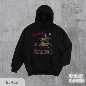 May include: Black hooded sweatshirt with a graphic print of a cowboy riding a horse with the text "Cowboy Rodeo" in red and blue.