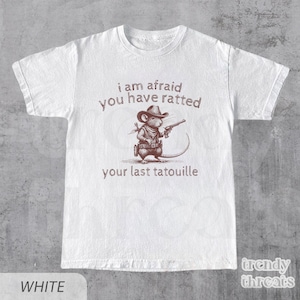 Pode incluir: Camiseta branca com um rato de desenho animado marrom usando um chap&eacute;u de cowboy e segurando uma arma. O texto na camiseta diz "i am afraid you have ratted your last tatouille".
