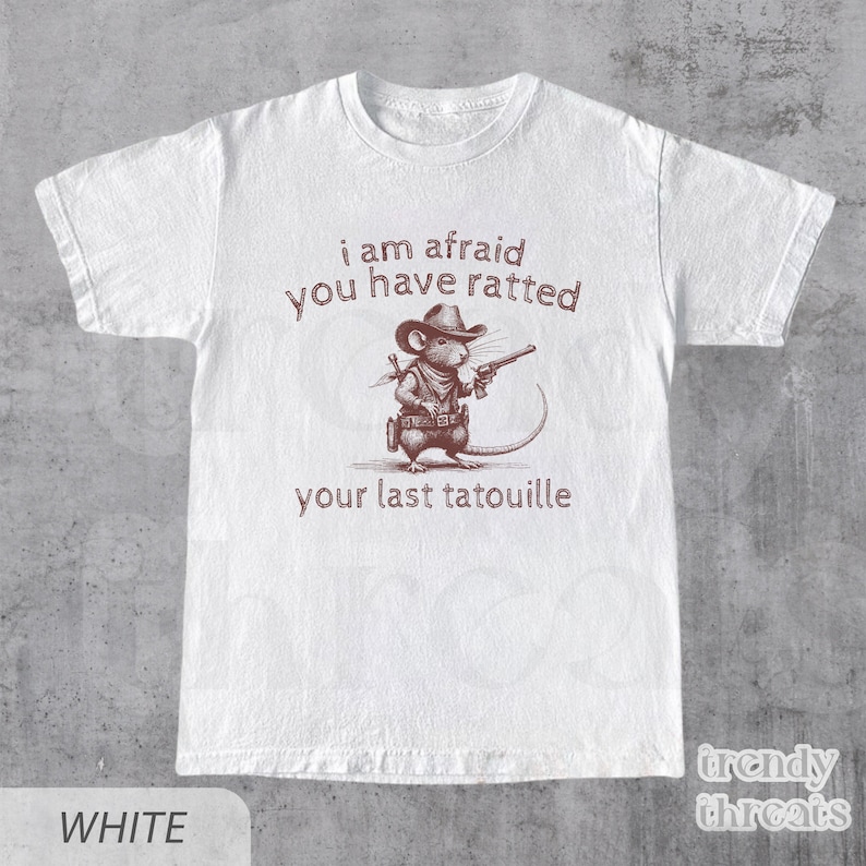 Pode incluir: Camiseta branca com um rato de desenho animado marrom usando um chap&eacute;u de cowboy e segurando uma arma. O texto na camiseta diz "i am afraid you have ratted your last tatouille".