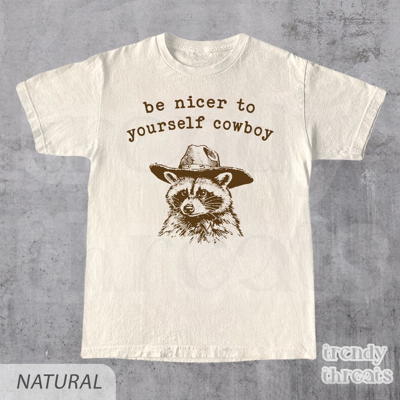 May include: A beige t-shirt with a brown raccoon wearing a cowboy hat and the text "be nicer to yourself cowboy".