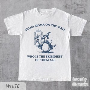 May include: White t-shirt featuring a blue illustration of a penguin wearing a wizard hat and holding a wand. The text "Sigma Sigma on the wall" and "Who is the Skibidiest of them all" is printed on the shirt.