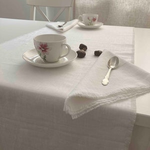 Könnte beinhalten: Ein weißer Tischläufer aus Leinen mit ausgefransten Kanten, eine weiße Teetasse aus Keramik mit Blumenmuster, ein silberner Löffel und ein paar Eicheln auf einem weißen Tisch.