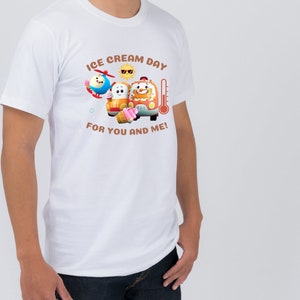 May include: White t-shirt with a cartoon graphic of a bus, a helicopter, and a sun wearing sunglasses. The text on the shirt reads "Ice Cream Day for You and Me!"
