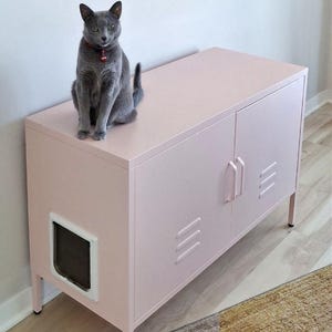 May include: A light pink metal cabinet with two doors featuring vertical ventilation slats and matching handles. A white pet door is installed on the left side panel. A grey cat with a red collar sits on the flat top surface of the cabinet.