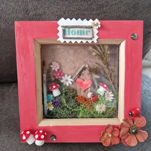 May include: A red framed shadow box featuring a miniature fairy garden scene. The scene includes a small house with a heart, mushrooms, flowers, and the word "Home". Decorative flowers and ladybirds are attached to the frame.
