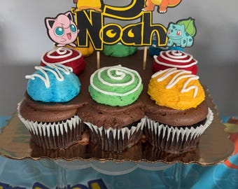 May include: A chocolate cupcake with blue, green, and orange frosting decorated with a Pokemon themed cake topper. The topper features a number 5, the name Noah, and various Pokemon characters including Pikachu, Bulbasaur, and Charmander.