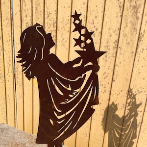 Könnte beinhalten: Eine rostige Metallsilhouette einer Person, die einen Stern hält. Die Person trägt eine lange, fließende Robe und hat den Kopf nach hinten geneigt. Die Silhouette steht auf einem runden Sockel.