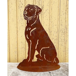 Könnte beinhalten: Rostbraune Metall-Hunde-Silhouette-Skulptur. Der Hund sitzt aufrecht, den Kopf leicht zur Seite geneigt. Die Skulptur ist auf einem kreisförmigen Sockel montiert. Der Hintergrund ist eine hellgelbe Holzwand.