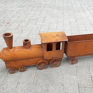Könnte beinhalten: Ein rostiger Metall-Zug-Pflanzkübel in Orangebraun. Der Zug besteht aus einer Lokomotive mit Schornstein, Führerhaus und einem separaten Wagen. Die Räder sind sichtbar. Maße in Zentimetern.