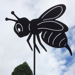 Peut inclure: Piquet de jardin en métal noir en forme de silhouette d'abeille. L'abeille est tournée vers la gauche et a un abdomen rayé. Le piquet est devant un arbre vert et un ciel bleu nuageux.