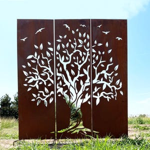 Op de afbeelding: Een metalen scherm met drie panelen met een boomontwerp. Het witte silhouet van de boom is uitgesneden uit de roestbruine panelen. Vogels zijn uit het metaal gesneden en lijken te vliegen. Het scherm staat buiten tegen een blauwe lucht.