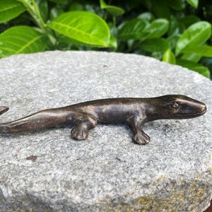Könnte beinhalten: Eine dunkel bronzefarbene Eidechsenfigur aus Metall mit einer glatten, polierten Oberfläche. Die Eidechse ist in einer horizontalen Kriechposition dargestellt, mit einem leicht nach oben gebogenen Schwanz, vier Beinen und einem Kopf.