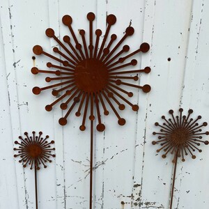 May include: Three rusty metal sculptures of stylised dandelions with long stems. The sculptures are arranged in a row against a white wooden background.