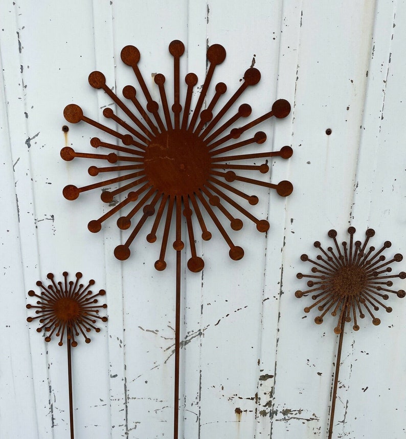 May include: Three rusty metal sculptures of stylised dandelions with long stems. The sculptures are arranged in a row against a white wooden background.