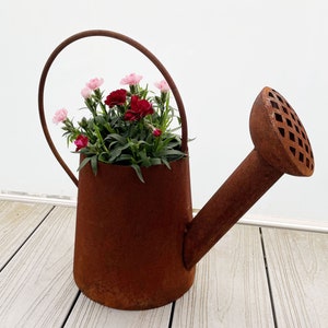 Könnte beinhalten: Eine rostige Gießkanne aus Metall mit einem breiten Ausguss und einem Griff. Die Kanne ist mit rosa und roten Nelken gefüllt.