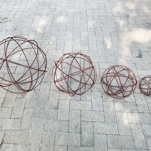 Op de afbeelding: Vier roestige metalen bollen van verschillende grootte, gerangschikt in een rij van groot naar klein. De bollen zijn gemaakt van draad en hebben een cirkelvormig ontwerp.