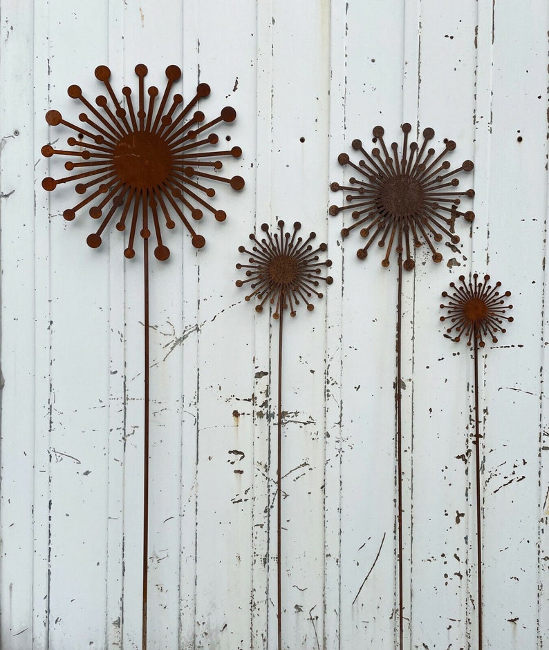 May include: Four rusty metal dandelion sculptures with long stems against a white painted wood background.