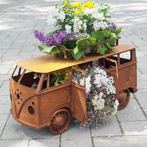 Op de afbeelding: Een roestbruine metalen Volkswagen bus bloembak gevuld met kleurrijke bloemen. De bus heeft een open deur en een bagagerek op het dak, overvol met paarse, witte en gele bloemen. De bloembak staat op een grijze stenen ondergrond.