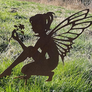 Può includere: Una silhouette di fata in metallo arrugginito con ali, seduta e che tiene un fiore. La fata si trova in un giardino con erba verde.