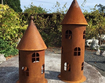 Könnte beinhalten: Zwei rostige Metall-Turm-Kerzenhalter mit einem spitzen Dach und ausgeschnittenen Fenstern. Der höhere Turm befindet sich rechts und der niedrigere links.