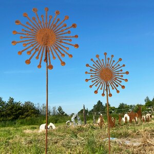 May include: Two rusty metal sculptures of stylised dandelions with a sunburst design. The sculptures are on tall metal poles and are set against a blue sky.