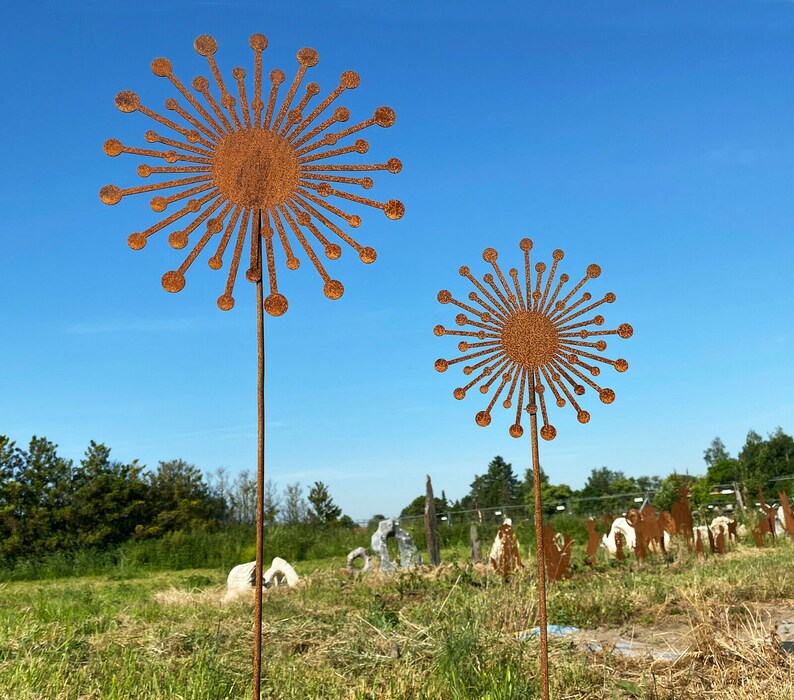 May include: Two rusty metal sculptures of stylised dandelions with a sunburst design. The sculptures are on tall metal poles and are set against a blue sky.