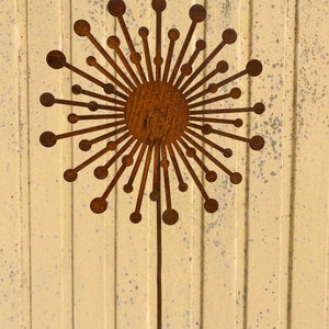 May include: A rusty brown metal dandelion sculpture with a round centre and many thin, pointed rays. The sculpture is mounted on a thin metal rod.