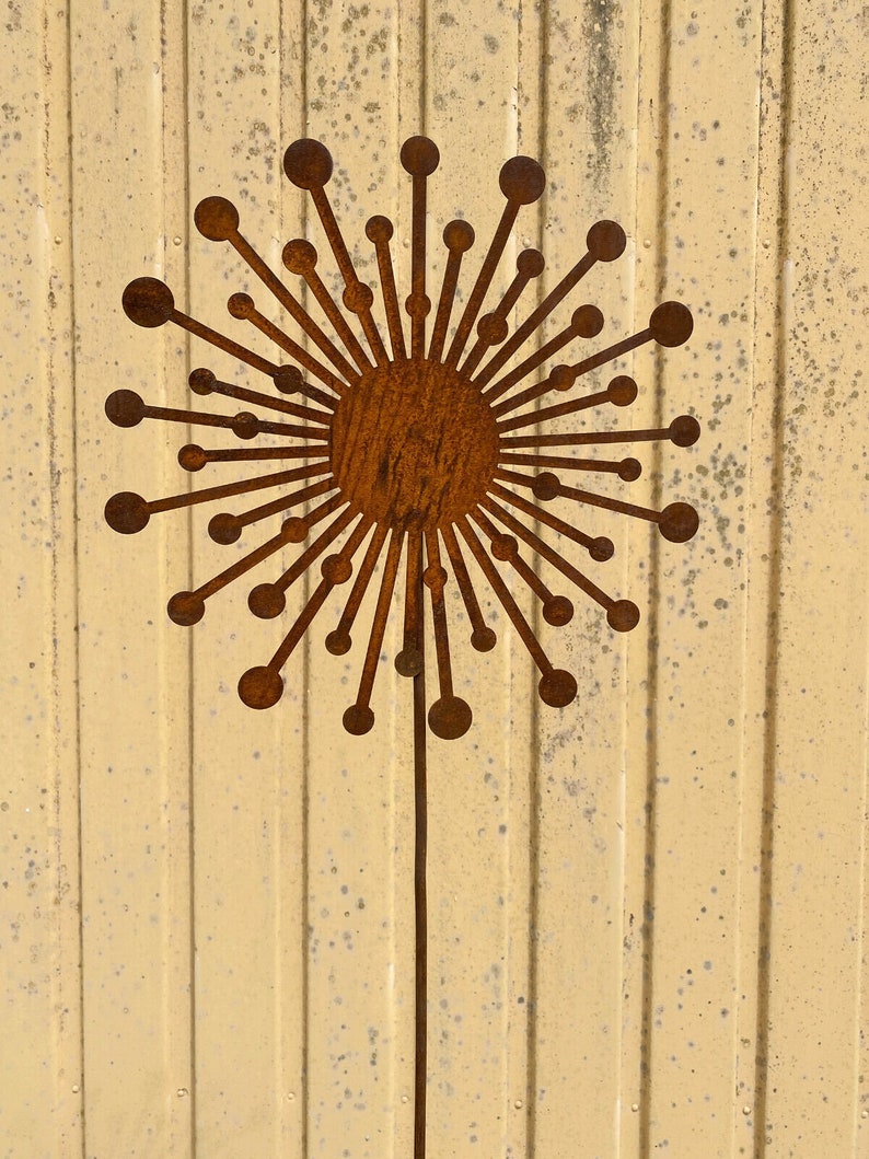 May include: A rusty brown metal dandelion sculpture with a round centre and many thin, pointed rays. The sculpture is mounted on a thin metal rod.