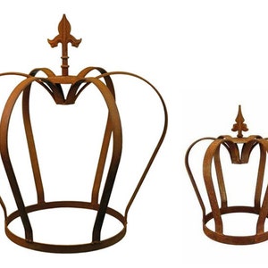 Op de afbeelding: Twee roestige metalen plantenstandaards in de vorm van een kroon. De grotere standaard is hoger en heeft een uitgebreider ontwerp. Beide standaards hebben een puntige top.