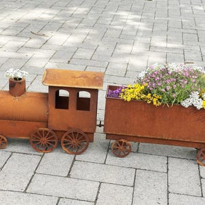 Puede incluir: Un jardinera con forma de tren de metal oxidado. La locomotora tiene una chimenea y una pequeña cabina. El vagón adjunto está lleno de flores coloridas, creando un encantador acento de jardín.