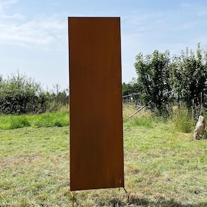Op de afbeelding: Een hoog, rechthoekig metalen paneel met een roestige afwerking. Het paneel staat op een grasveld met een blauwe lucht op de achtergrond.