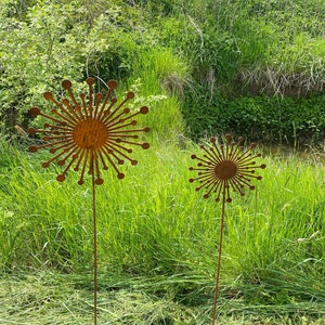 May include: Two rusty metal sculptures of stylised flowers with long stems stand in a grassy field. The flowers are made of thin metal and have a circular centre with radiating spokes.