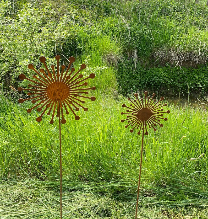 May include: Two rusty metal sculptures of stylised flowers with long stems stand in a grassy field. The flowers are made of thin metal and have a circular centre with radiating spokes.