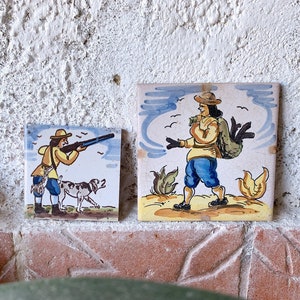 Könnte beinhalten: Zwei antike Keramikfliesen, die Szenen von Jägern mit Hunden darstellen. Die erste Fliese zeigt einen Jäger in einem gelben Hemd und blauen Hosen, der mit einer Waffe auf einen Vogel zielt, während sein Hund neben ihm läuft. Die zweite Fliese zeigt einen Jäger in einem blauen Hemd und blauen Hosen, der eine Tasche auf dem Rücken trägt und durch ein Feld geht.